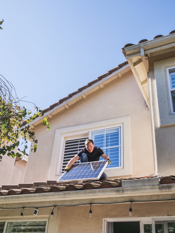 man-in-blue-shirt-and-black-pants-sitting-on-roof-9875407 A worker installs a solar panel on a residential rooftop, showcasing renewable energy technology.