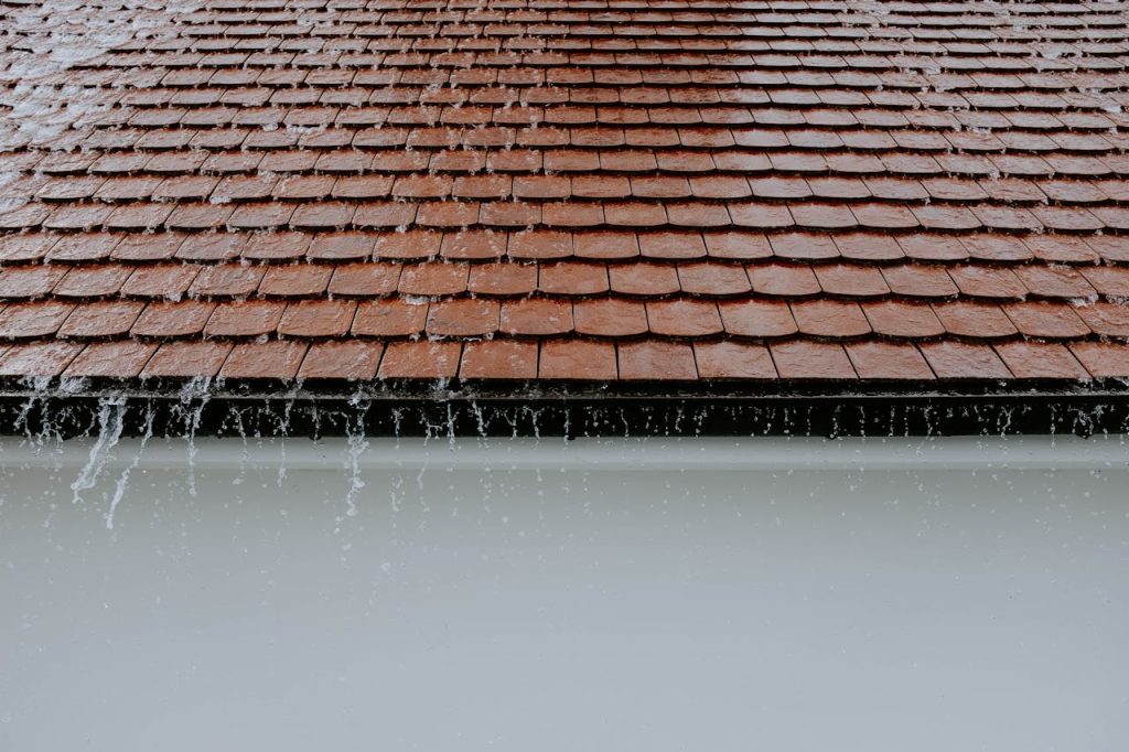 photo-of-roof-while-raining-2663254 Brown roof tiles with rainwater cascading down, showcasing architectural texture and detail.